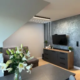 Nowoczesny salon z szarą ścianą dekoracyjną, podświetlaną listwami LED, ciemną komodą z drewnianym blatem, telewizorem, szarą sofą i stołem z bukietem białych lilii w wazonie.