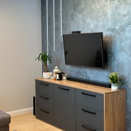 PRYIMENKO Sp. z o.o. - Szara komoda z drewnianym blatem, czarnymi uchwytami i sprzętem audio pod telewizorem na tle ściany z efektem betonu, oświetlenie LED w suficie.