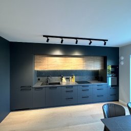 PRYIMENKO Sp. z o.o. - Nowoczesna kuchnia z szarymi szafkami i drewnianymi akcentami, czarny zlew i bateria, oświetlenie punktowe na suficie, widoczne elementy wyposażenia, takie jak noże i przyprawy.