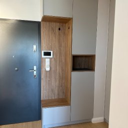 PRYIMENKO Sp. z o.o. - Szara szafa w przedpokoju z drewnianym panelem, wbudowane siedzisko, wieszaki i panel sterowania domem obok drzwi wejściowych.