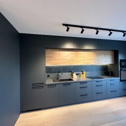 PRYIMENKO Sp. z o.o. - Nowoczesna kuchnia w odcieniach szarości z drewnianymi akcentami, oświetleniem podszafkowym LED i czarnymi uchwytami, widok na zabudowę kuchenną z blatem i sprzętami AGD.