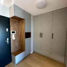 Szara zabudowa przedpokoju z drewnianym panelem z wieszakami i siedziskiem, obok drzwi wejściowe i szafa z czarnymi uchwytami, jasna podłoga.