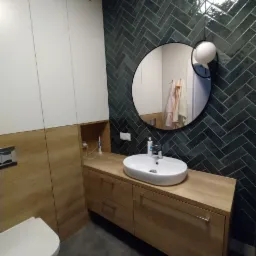 Nowoczesna łazienka z umywalką nablatową na drewnianej szafce, ciemnozielone płytki ułożone w jodełkę na ścianie, okrągłe lustro i minimalistyczna lampa wisząca.