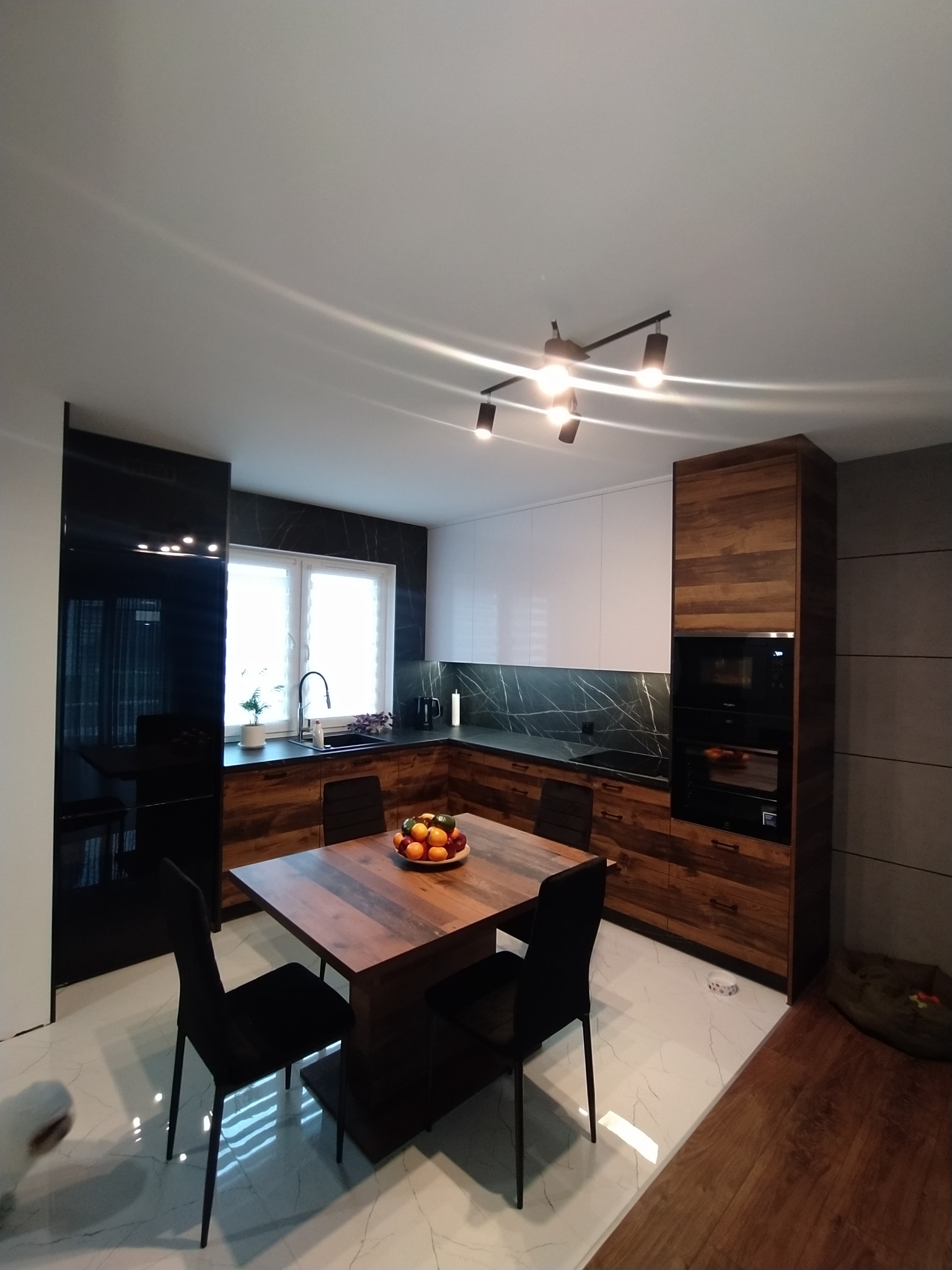 Nowoczesna kuchnia z drewnianymi frontami, białymi szafkami górnymi i czarnym panelem nad blatem, stół z owocami i czarne krzesła, oświetlenie punktowe.