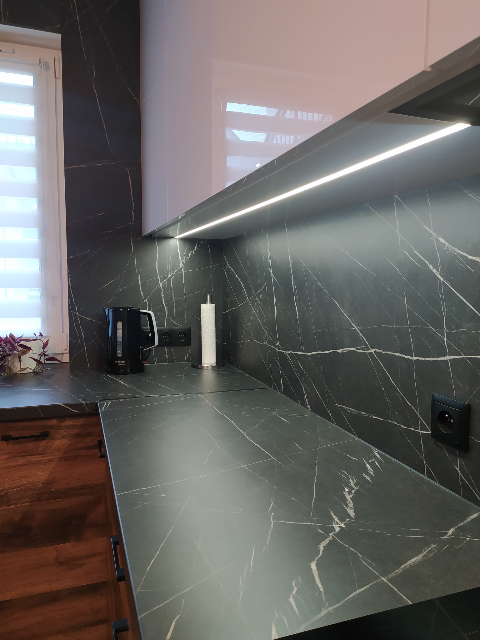 Fragment nowoczesnej kuchni z blatem imitującym czarny marmur z białymi żyłami, podświetlonym oświetleniem LED podszafkowym. Na blacie czajnik elektryczny Bosch, pojemnik na ręczniki papierowe...