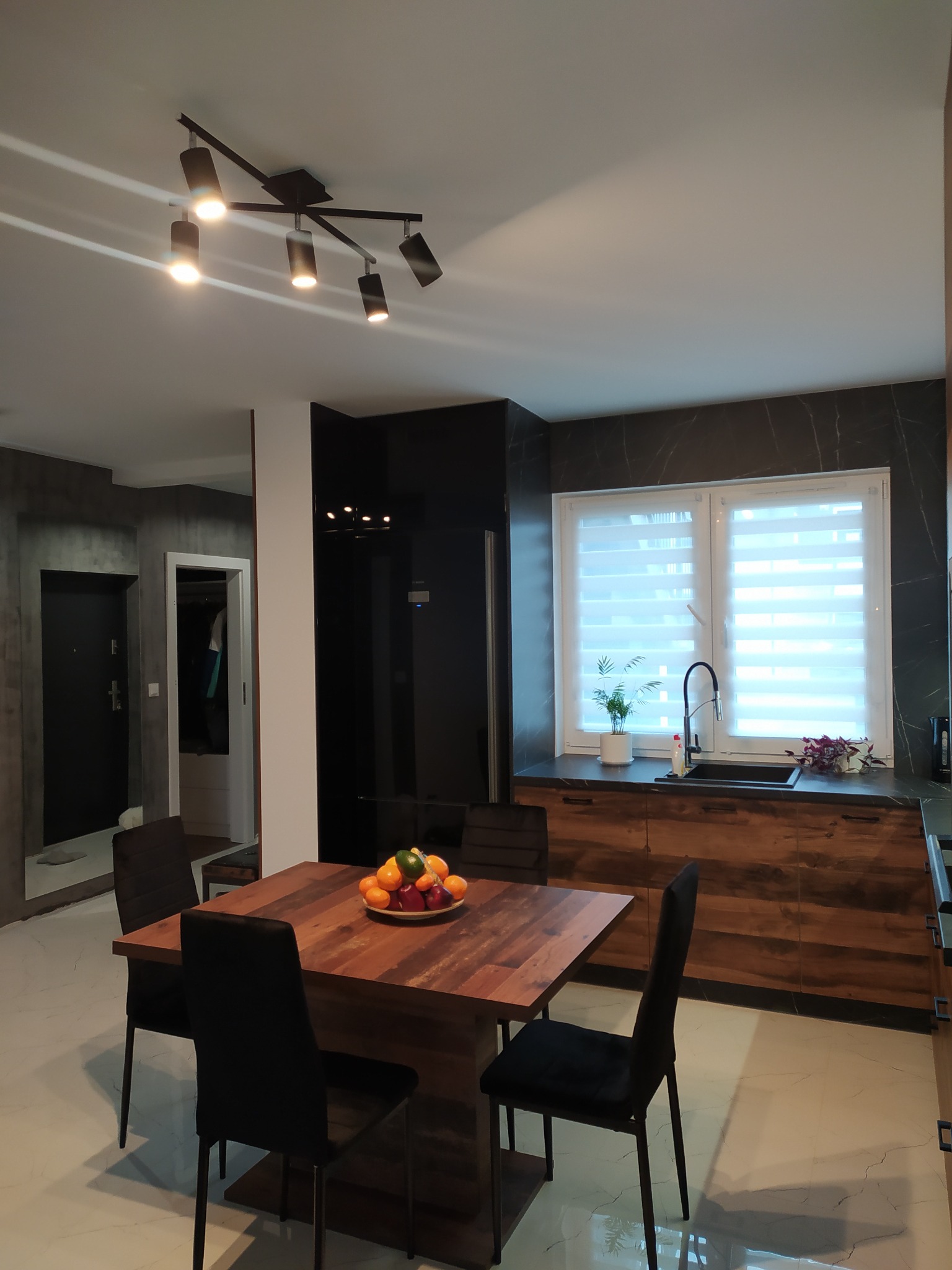 Nowoczesna kuchnia z drewnianymi szafkami, czarnym blatem i marmurową ścianą, oświetlona reflektorami sufitowymi; na stole misa z owocami.