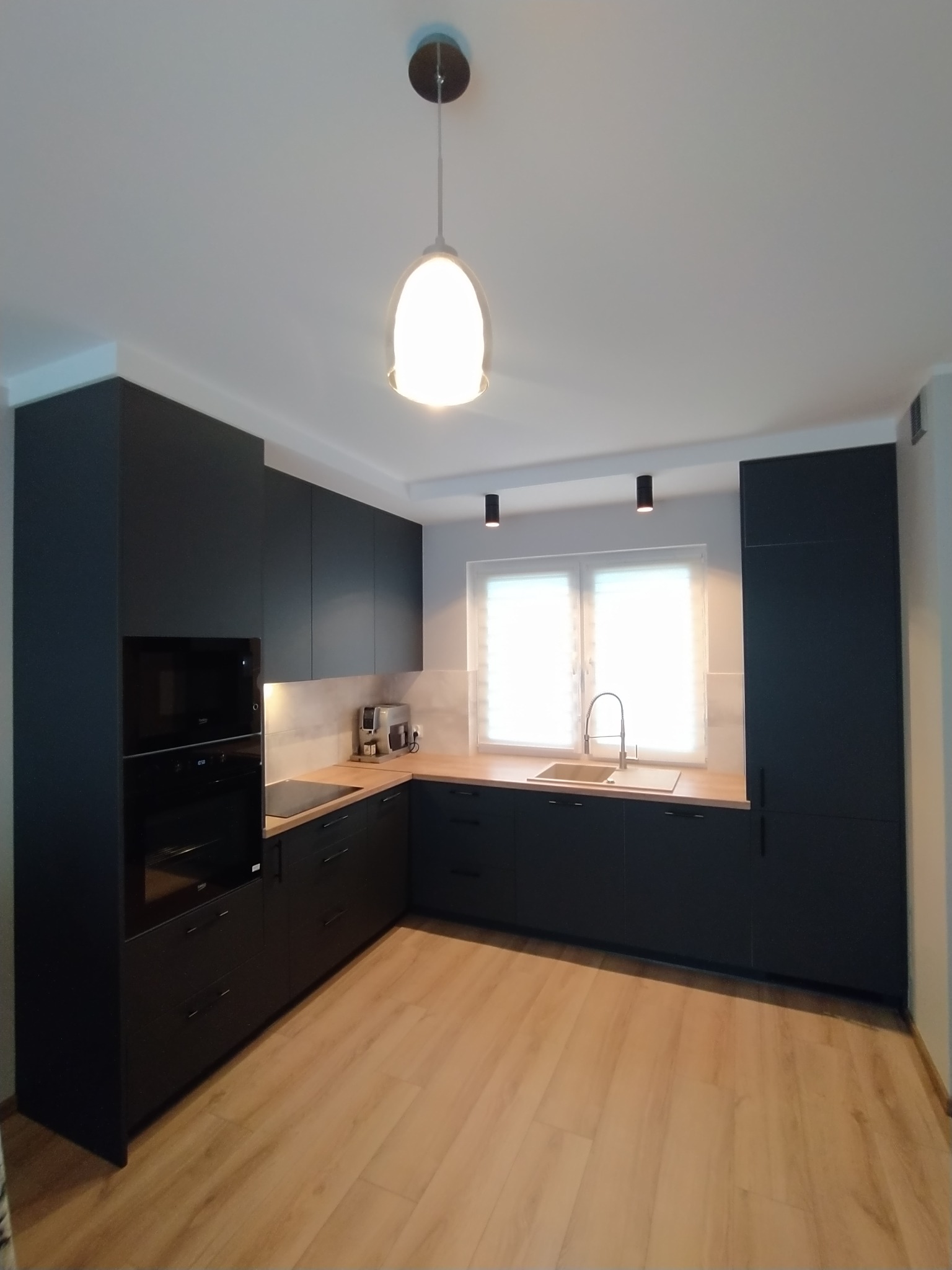 Nowoczesna kuchnia z czarnymi matowymi frontami, drewnianym blatem i jasną podłogą, oświetlona lampą wiszącą i spotami sufitowymi.