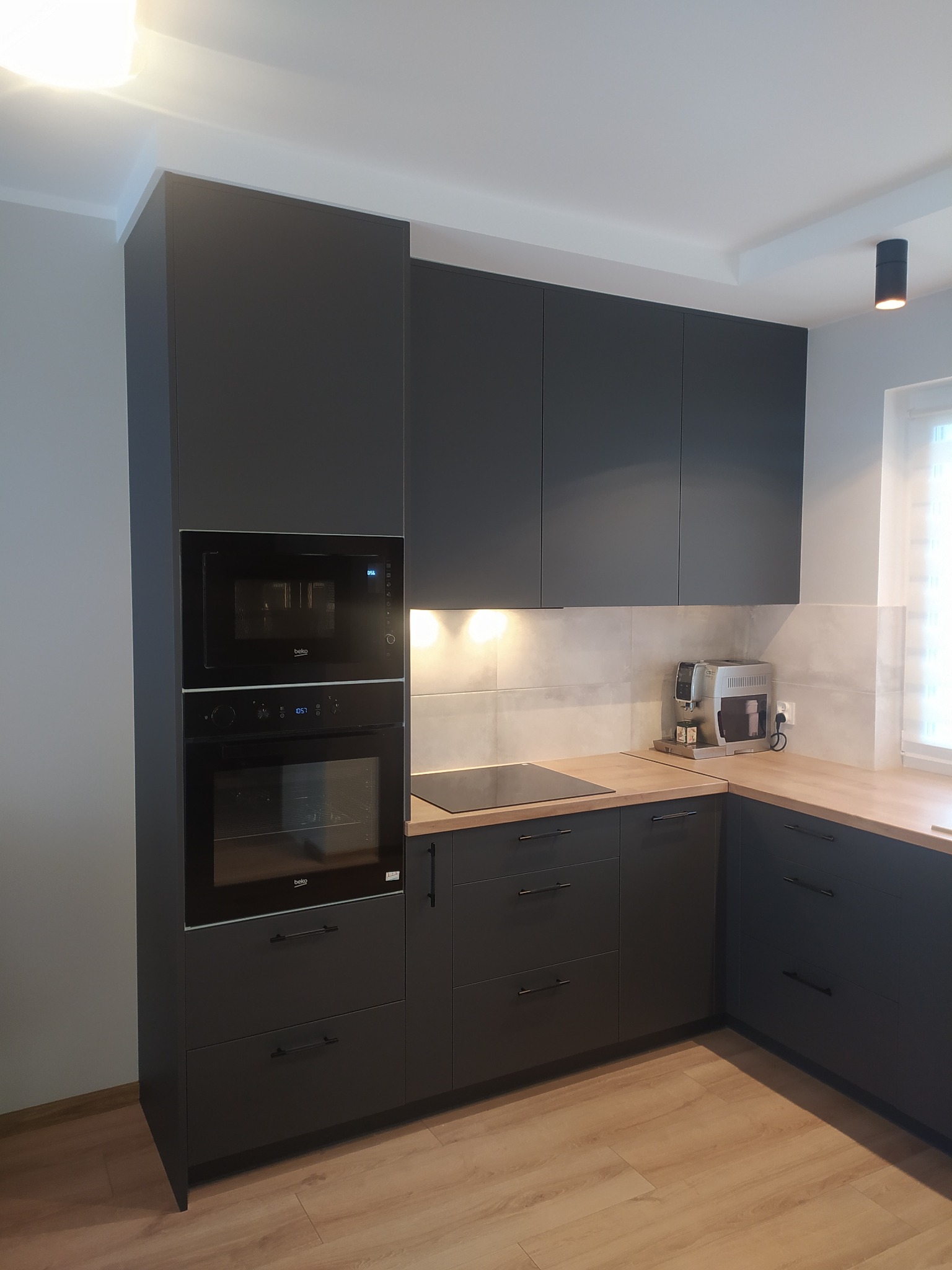 Nowoczesna kuchnia w odcieniach grafitu z drewnianym blatem, zabudowanymi piekarnikiem i mikrofalówką marki Beko oraz ekspres do kawy na blacie.