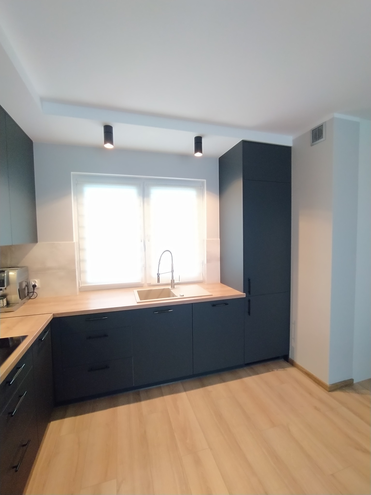 Nowoczesna kuchnia z ciemnymi, matowymi frontami szafek, jasnym blatem drewnianym i minimalistyczną armaturą, oświetlona dwoma punktowymi lampami sufitowymi.