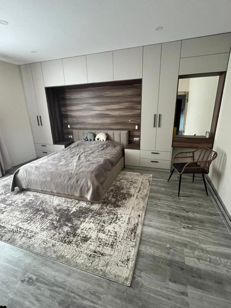Sypialnia z zabudową na wymiar: jasne szafy zintegrowane z wezgłowiem łóżka z drewnianym panelem, dywan w odcieniach szarości, krzesło wiklinowe i toaletka z lustrem.