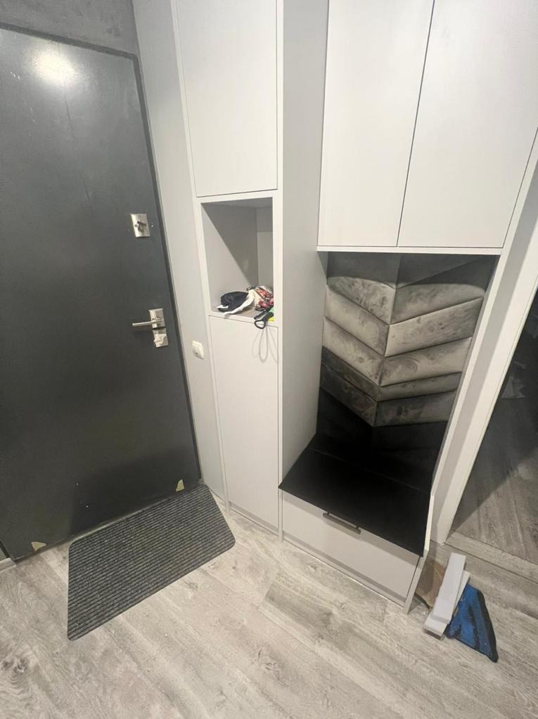 Wnęka z siedziskiem wykończona tapicerowanym panelem w kształcie chevronu, część zabudowy meblowej przedpokoju z szafkami i otwartą półką, widoczna czarna wycieraczka i drzwi wejściowe.