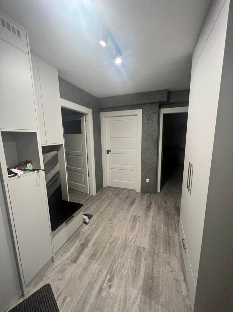 Przedpokój z szarą podłogą imitującą drewno, białą zabudową meblową z szafkami i siedziskiem, oraz dwiema białymi drzwiami na tle szarej ściany z dekoracyjnym wykończeniem.