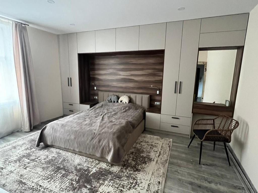 Szafa wnękowa z zabudową łóżka: jasne fronty, drewniana wnęka nad wezgłowiem, szara narzuta na łóżku, dywan w odcieniach szarości, krzesło z rattanu.