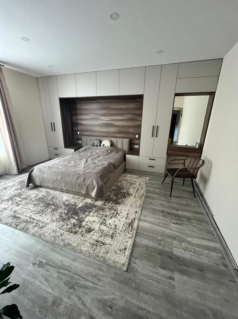 Sypialnia z łóżkiem, szarą pościelą i dekoracyjnymi poduszkami w kształcie słoni, zabudową meblową z szafami i drewnianym panelem za wezgłowiem, dywanem w odcieniach szarości i beżu oraz rattanowym...