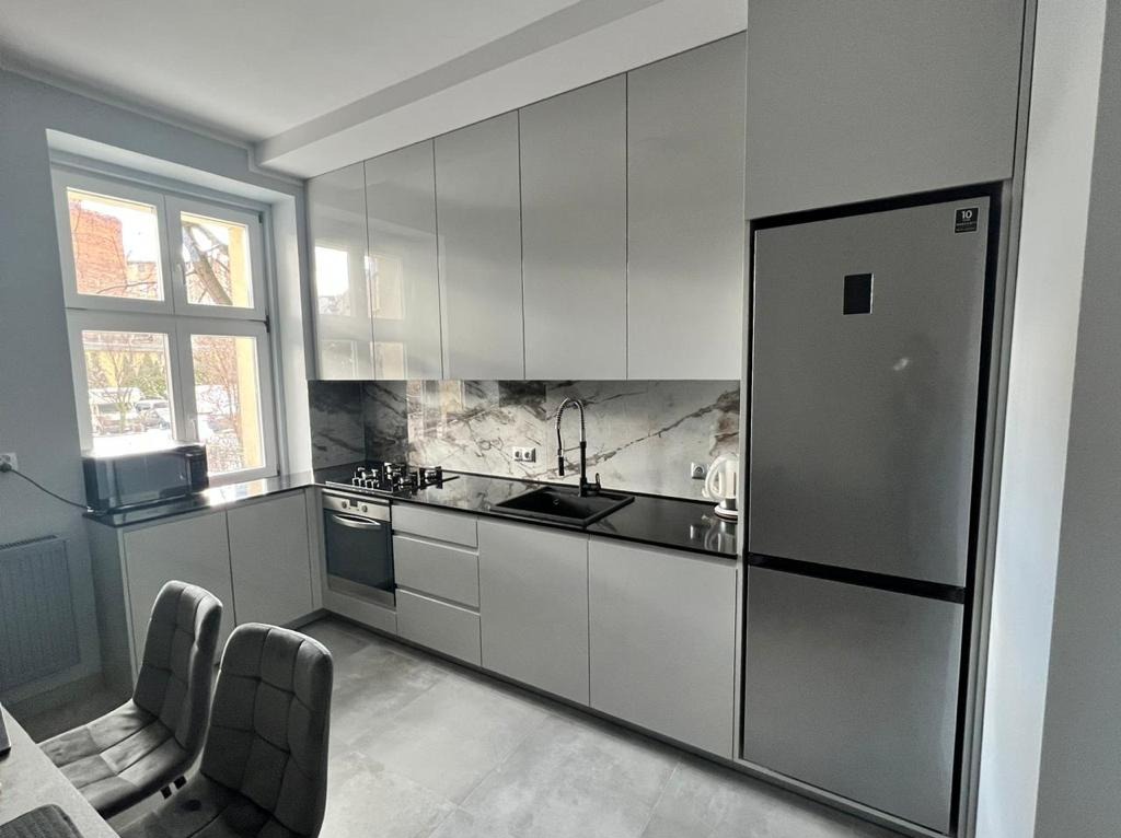 Nowoczesna kuchnia z szarymi, lakierowanymi frontami, czarnym blatem i marmurowym backsplashiem, oświetlona naturalnym światłem z okna.