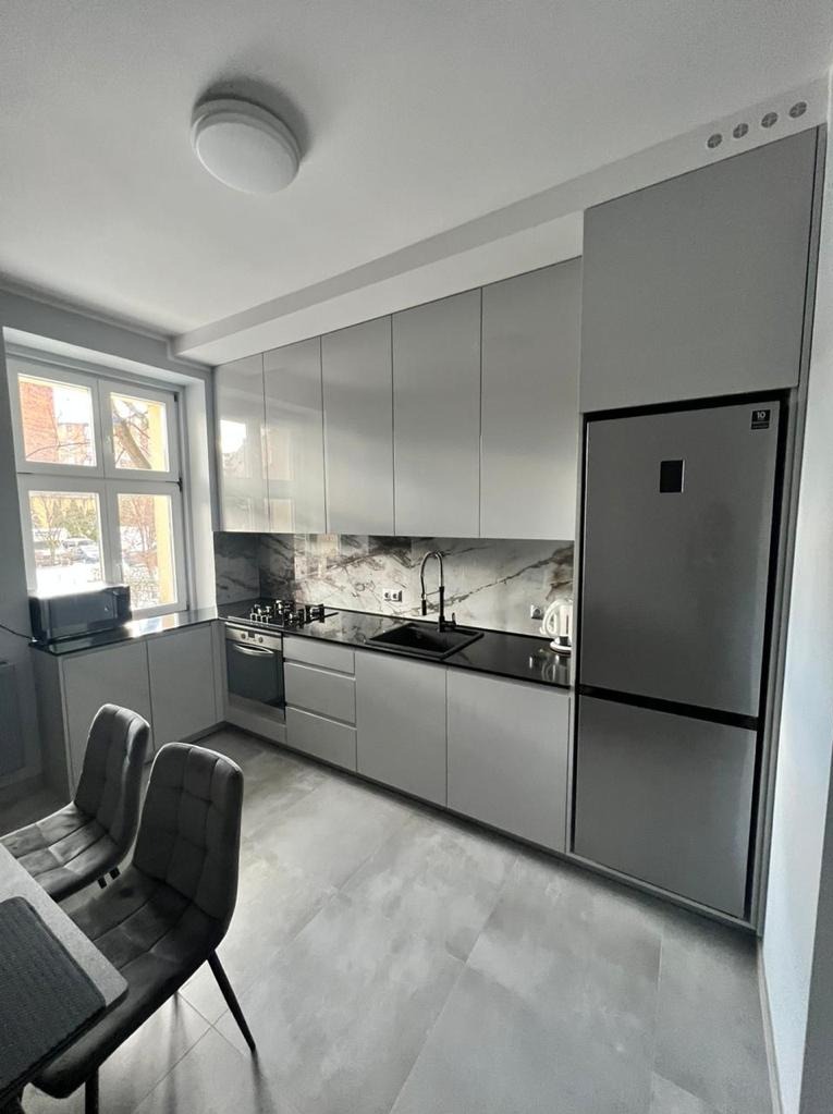 Nowoczesna kuchnia w odcieniach szarości z błyszczącymi frontami szafek, czarnym blatem i marmurowym backsplashiem. Widoczne krzesła przy stole, okno z widokiem na zewnątrz oraz wbudowana lodówka.