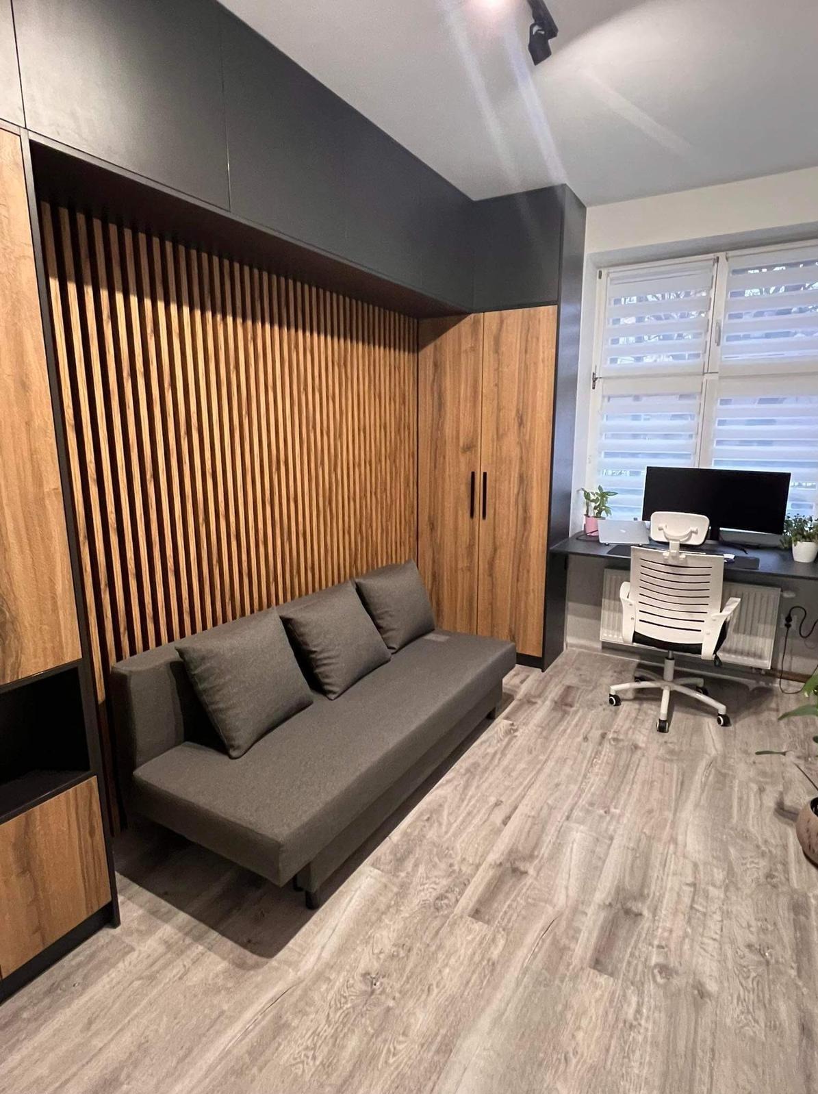 Pokój z szarą sofą z trzema poduszkami, zabudową meblową z drewnianymi lamelami i szafą z frontami w kolorze drewna, biurkiem i krzesłem biurowym pod oknem.