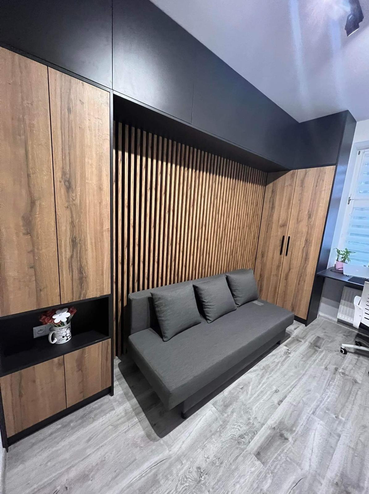 Szara rozkładana sofa z trzema poduszkami wkomponowana w zabudowę meblową z drewnopodobnymi frontami i czarnymi akcentami, widoczne pionowe listwy dekoracyjne na ścianie.