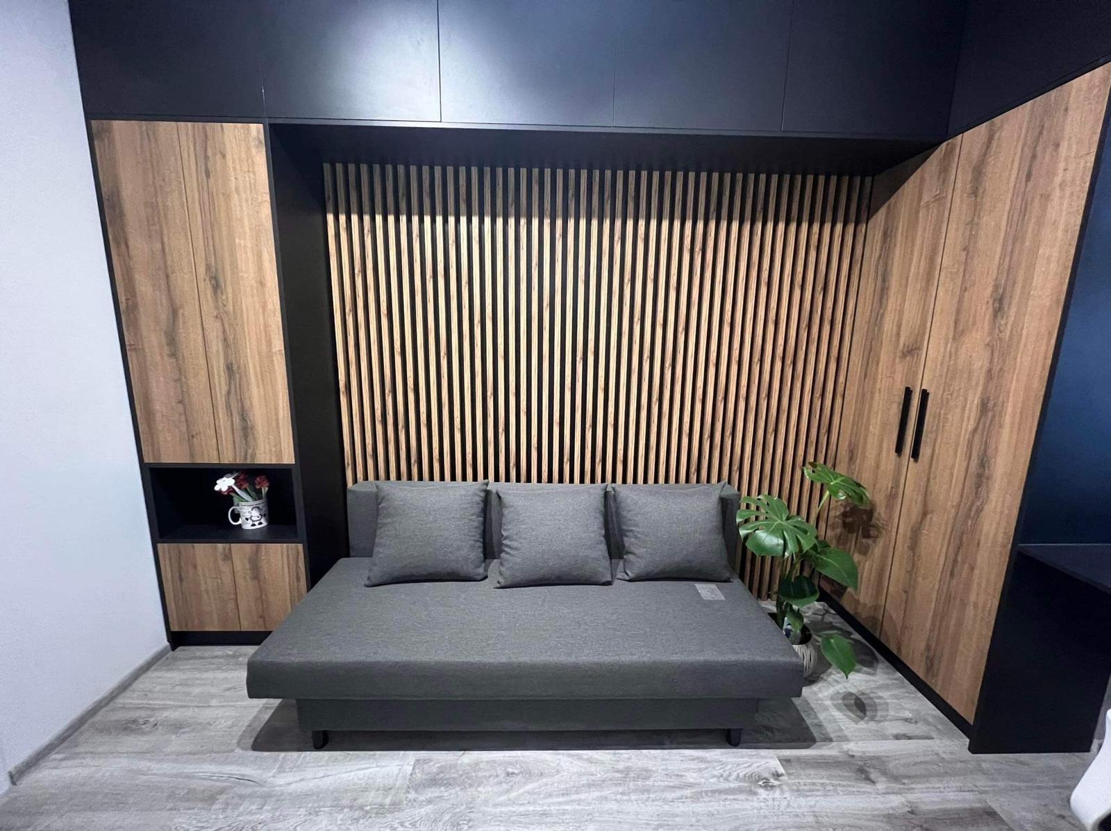 Szara sofa z trzema poduszkami na tle drewnianej ściany z pionowych listew i szafy z drewnianymi frontami, całość w nowoczesnym wnętrzu.