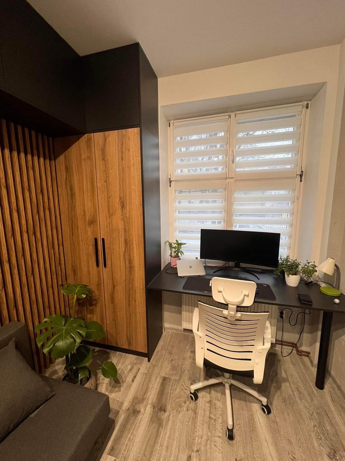 Nowoczesne biuro domowe z drewnianą szafą na wymiar z pionowymi lamelami, czarnym blatem biurka, białym krzesłem ergonomicznym, komputerem i roślinami doniczkowymi przy oknie z żaluzjami.