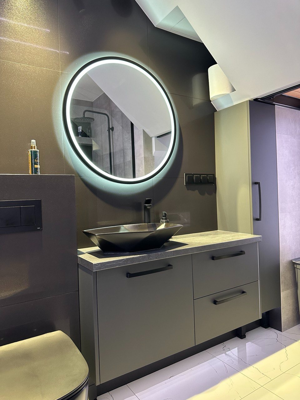 Nowoczesna łazienka w odcieniach szarości z okrągłym lustrem LED, czarną umywalką nablatową o geometrycznym kształcie i szafką z szufladami. W tle widoczny fragment kabiny prysznicowej.