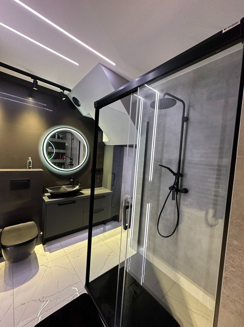 Nowoczesna łazienka w odcieniach szarości z czarnymi akcentami, kabina prysznicowa typu walk-in z czarną armaturą, umywalka nablatowa na szafce z szufladami, okrągłe lustro z podświetleniem LED...