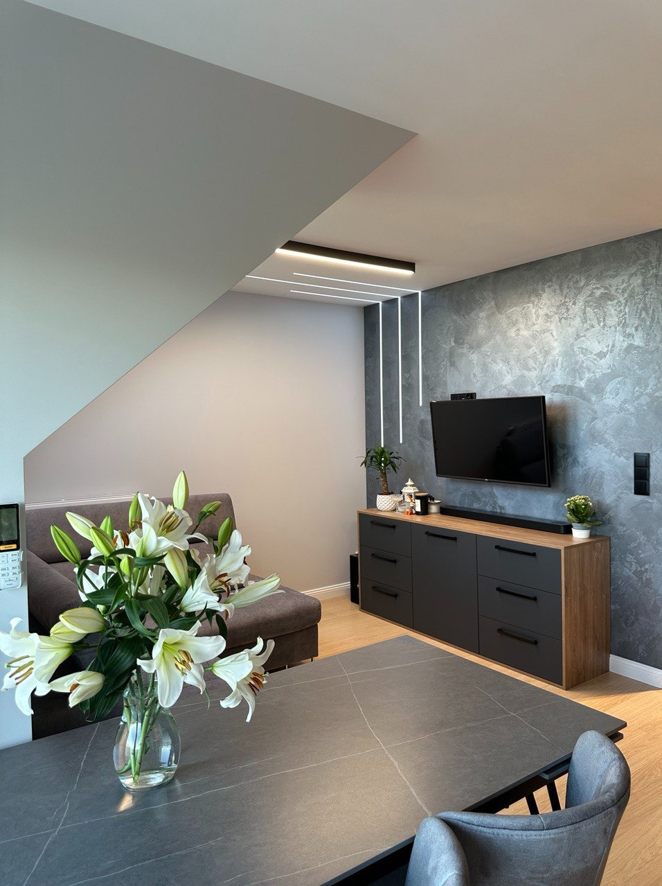 Nowoczesny salon z szarą ścianą dekoracyjną, podświetlaną listwami LED, ciemną komodą z drewnianym blatem, telewizorem, szarą sofą i stołem z bukietem białych lilii w wazonie.