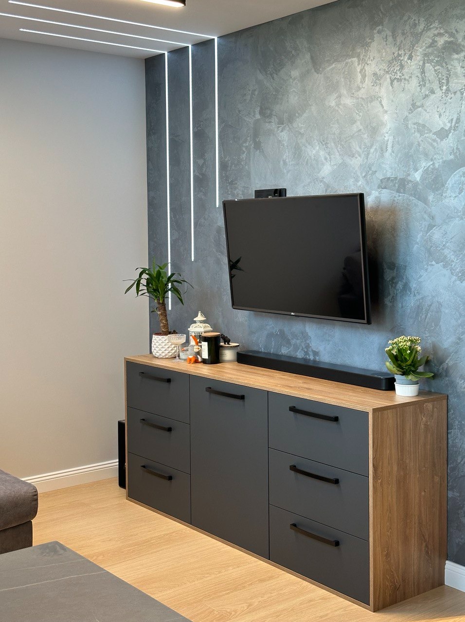 Szara komoda z drewnianym blatem, czarnymi uchwytami i sprzętem audio pod telewizorem na tle ściany z efektem betonu, oświetlenie LED w suficie.
