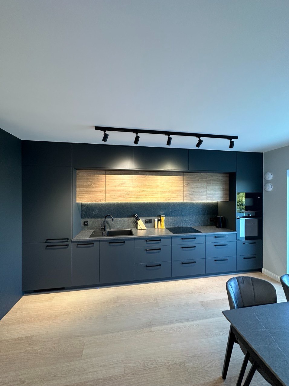 Nowoczesna kuchnia z szarymi szafkami i drewnianymi akcentami, czarny zlew i bateria, oświetlenie punktowe na suficie, widoczne elementy wyposażenia, takie jak noże i przyprawy.