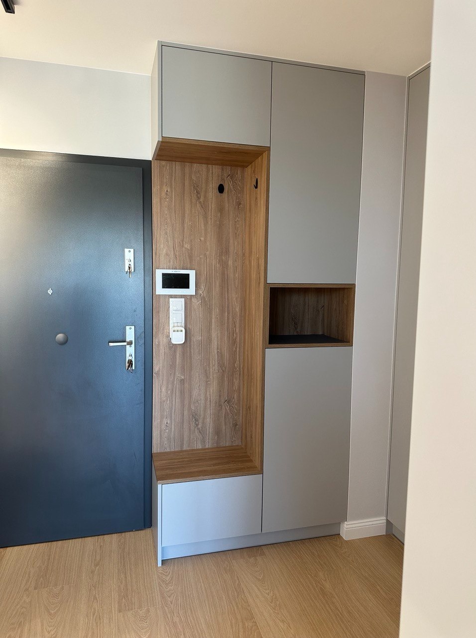 Szara szafa w przedpokoju z drewnianym panelem, wbudowane siedzisko, wieszaki i panel sterowania domem obok drzwi wejściowych.