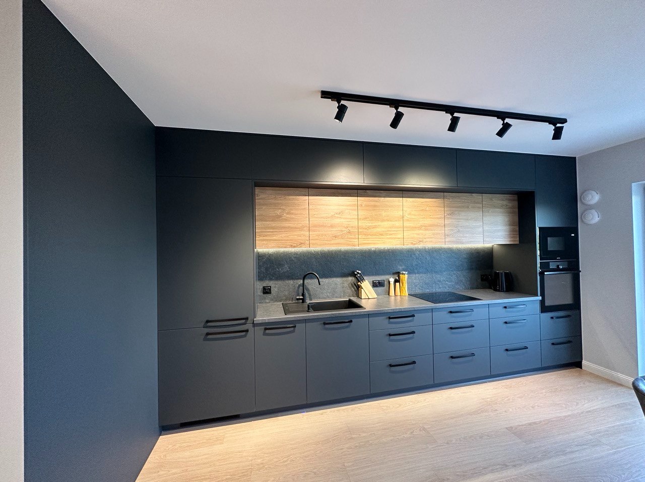 Nowoczesna kuchnia w odcieniach szarości z drewnianymi akcentami, oświetleniem podszafkowym LED i czarnymi uchwytami, widok na zabudowę kuchenną z blatem i sprzętami AGD.