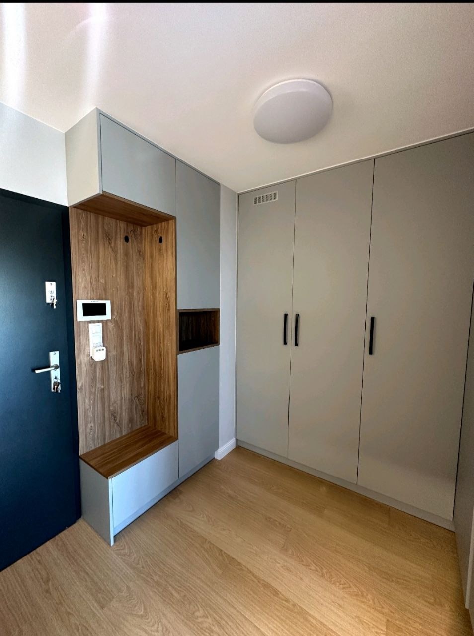 Szara zabudowa przedpokoju z drewnianym panelem z wieszakami i siedziskiem, obok drzwi wejściowe i szafa z czarnymi uchwytami, jasna podłoga.