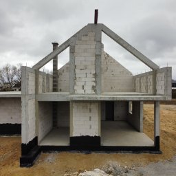 DEFTO MICHAŁ FORTUŃSKI - Surowy stan domu z białego pustaka, widoczne fundamenty z izolacją, komin i konstrukcja dachu. Na zewnątrz piach i częściowo ułożone deski.