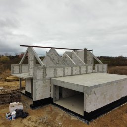 DEFTO MICHAŁ FORTUŃSKI - Surowy stan domu z garażem w budowie, ściany z białego bloczka, widoczne palety z materiałami, przenośna toaleta w tle, pochmurne niebo.