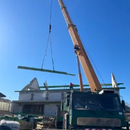 Żuraw budowlany podnosi belkę na budowę domu z białego pustaka. Widoczny pracownik na dachu i materiały budowlane. Słoneczny dzień, niebieskie niebo.