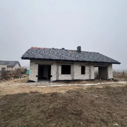 Dom w trakcie budowy, ściany z białego bloczka, dach z czarnej dachówki. Widoczne okna i komin. Szary, pochmurny dzień. Nierówny teren wokół budynku.