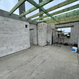 Wnętrze domu w trakcie budowy: ściany z betonu komórkowego, strop z drewnianych belek pomalowanych na zielono, rusztowanie przy oknie, betonowa podłoga.