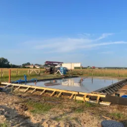 Wylany fundament pod budowę domu jednorodzinnego. Widoczna drewniana szalówka, zbrojenie i świeży beton. W tle maszyna do betonu i zielone pola.