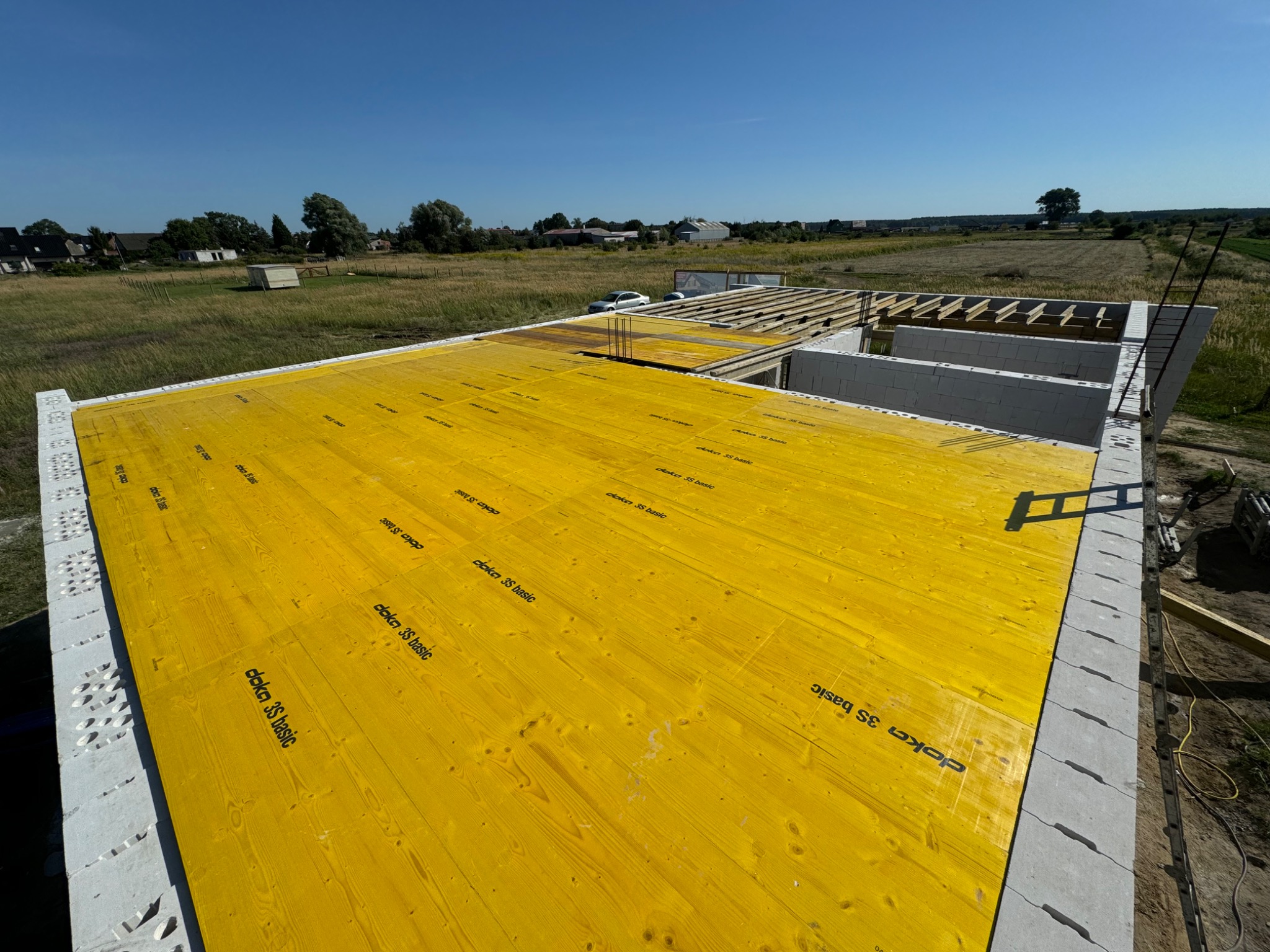 Budowa domu jednorodzinnego: widok z góry na fundamenty i ściany z bloczków, żółte płyty Doka 3S basic na podłodze, widoczny szkielet dachu i krajobraz w tle.