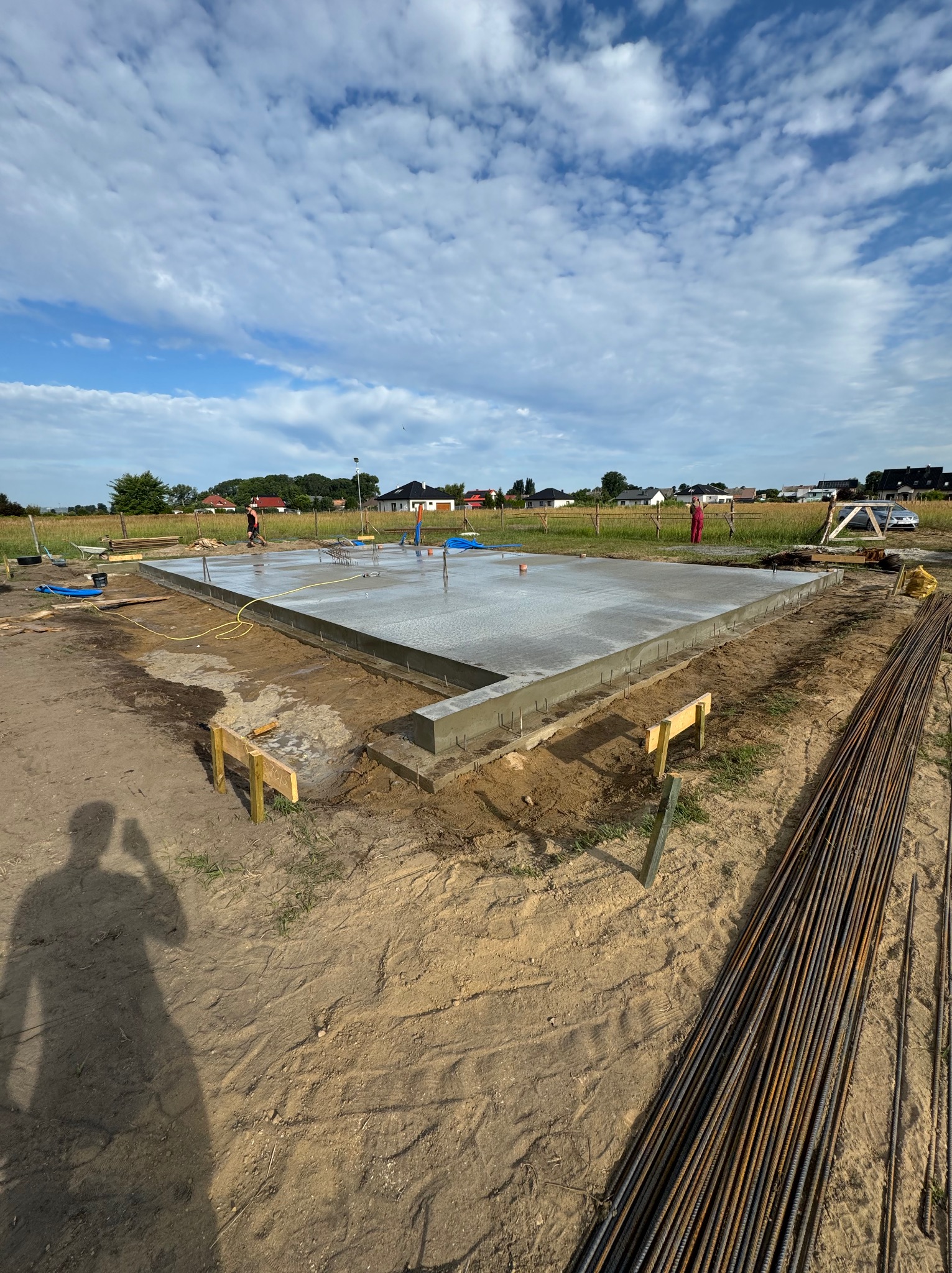 Wylany fundament z wystającymi prętami zbrojeniowymi, widok z poziomu gruntu z shadow-selfie fotografa, w tle domy jednorodzinne i błękitne niebo z obłokami.