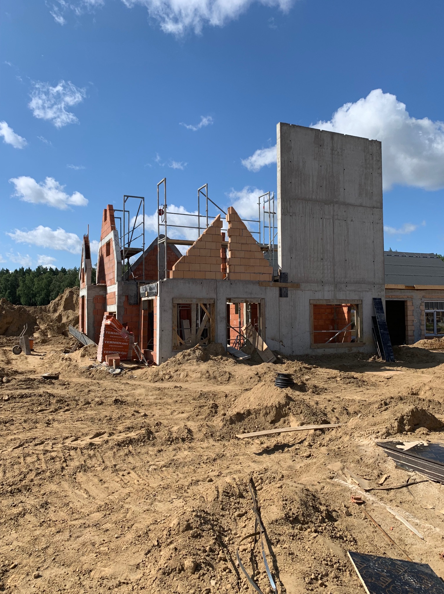 Budowa domu murowanego w trakcie realizacji: widoczne ściany z czerwonej cegły, betonowy szkielet, rusztowania i hałdy piasku na placu budowy w słoneczny dzień.