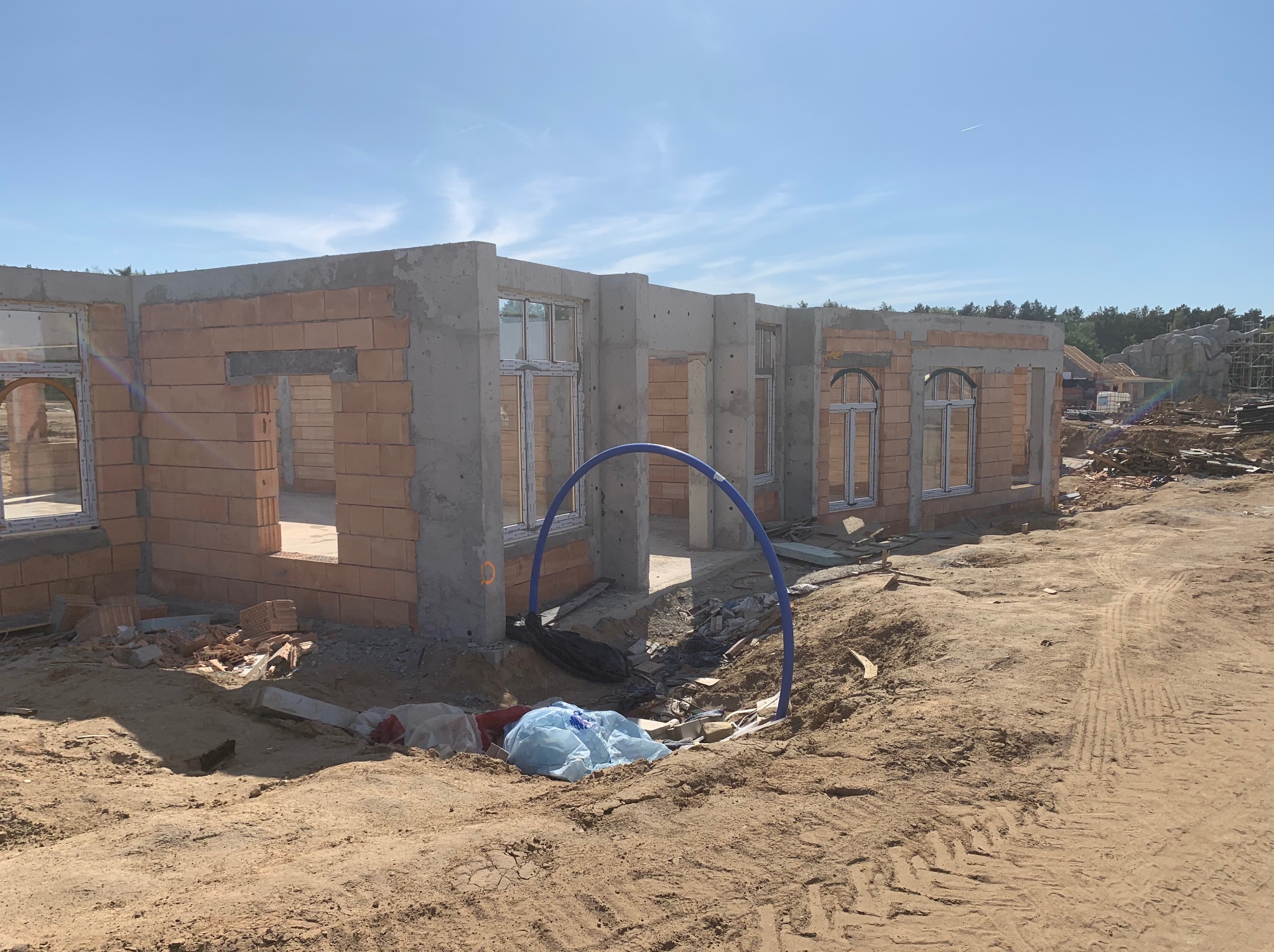 Surowy stan budynku z czerwonej cegły i betonu, widoczne otwory okienne, niebieskie rury instalacyjne na placu budowy, ziemia z śladami opon.