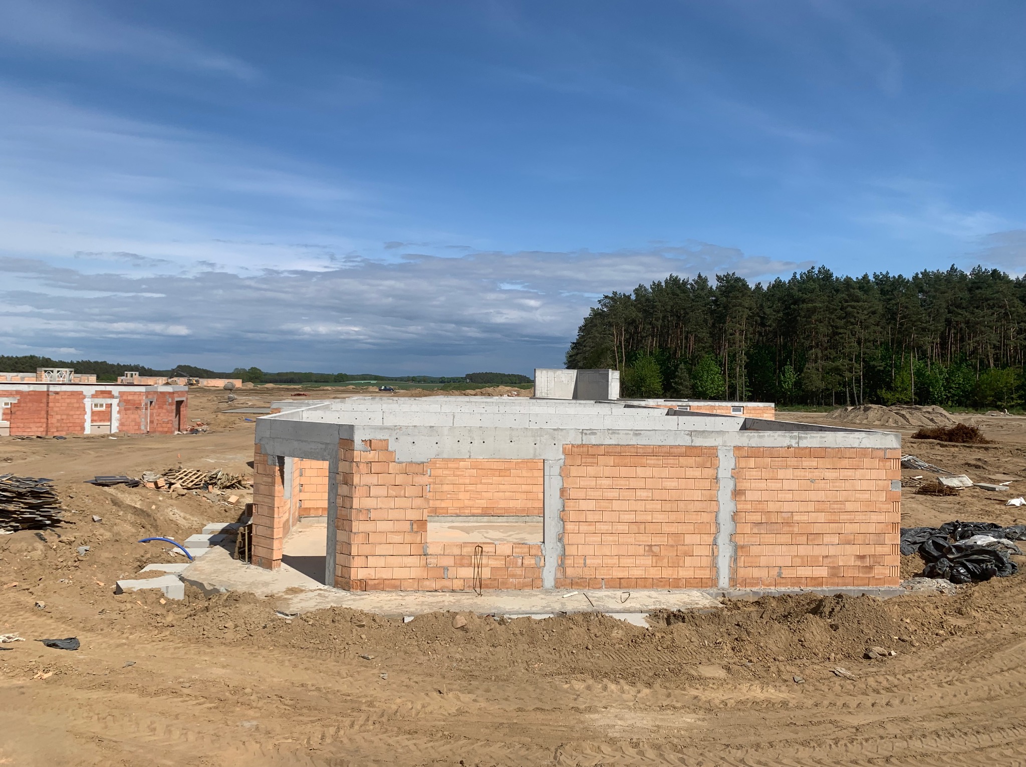 Budowa domu jednorodzinnego w Kliniskach Wielkich, widoczne ściany z czerwonej cegły silikatowej i betonowe elementy konstrukcyjne, stan surowy otwarty, w tle las.