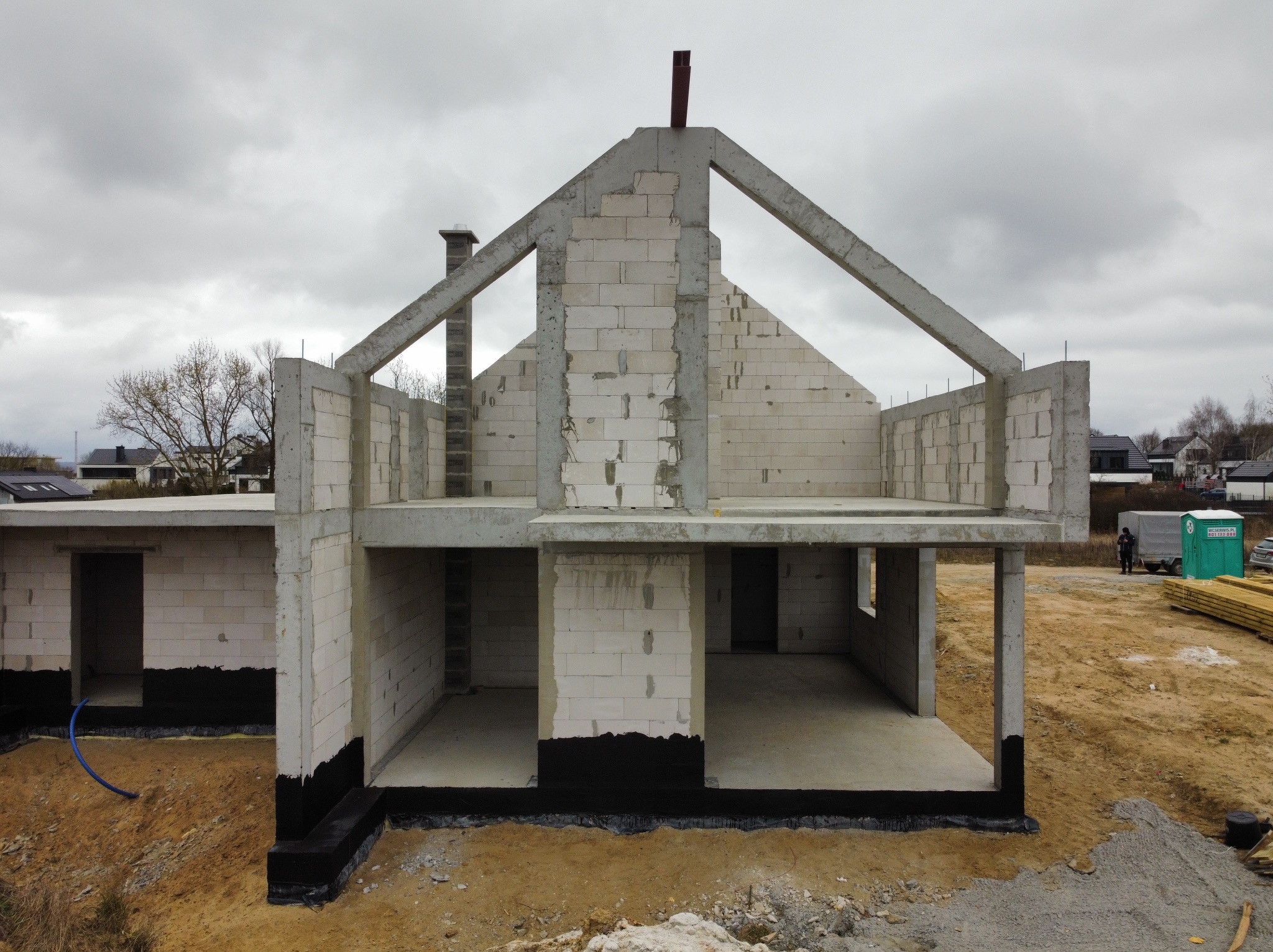 Surowy stan domu z białego pustaka, widoczne fundamenty z izolacją, komin i konstrukcja dachu. Na zewnątrz piach i częściowo ułożone deski.
