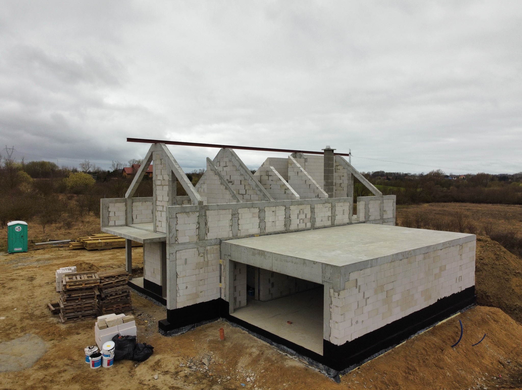 Surowy stan domu z garażem w budowie, ściany z białego bloczka, widoczne palety z materiałami, przenośna toaleta w tle, pochmurne niebo.