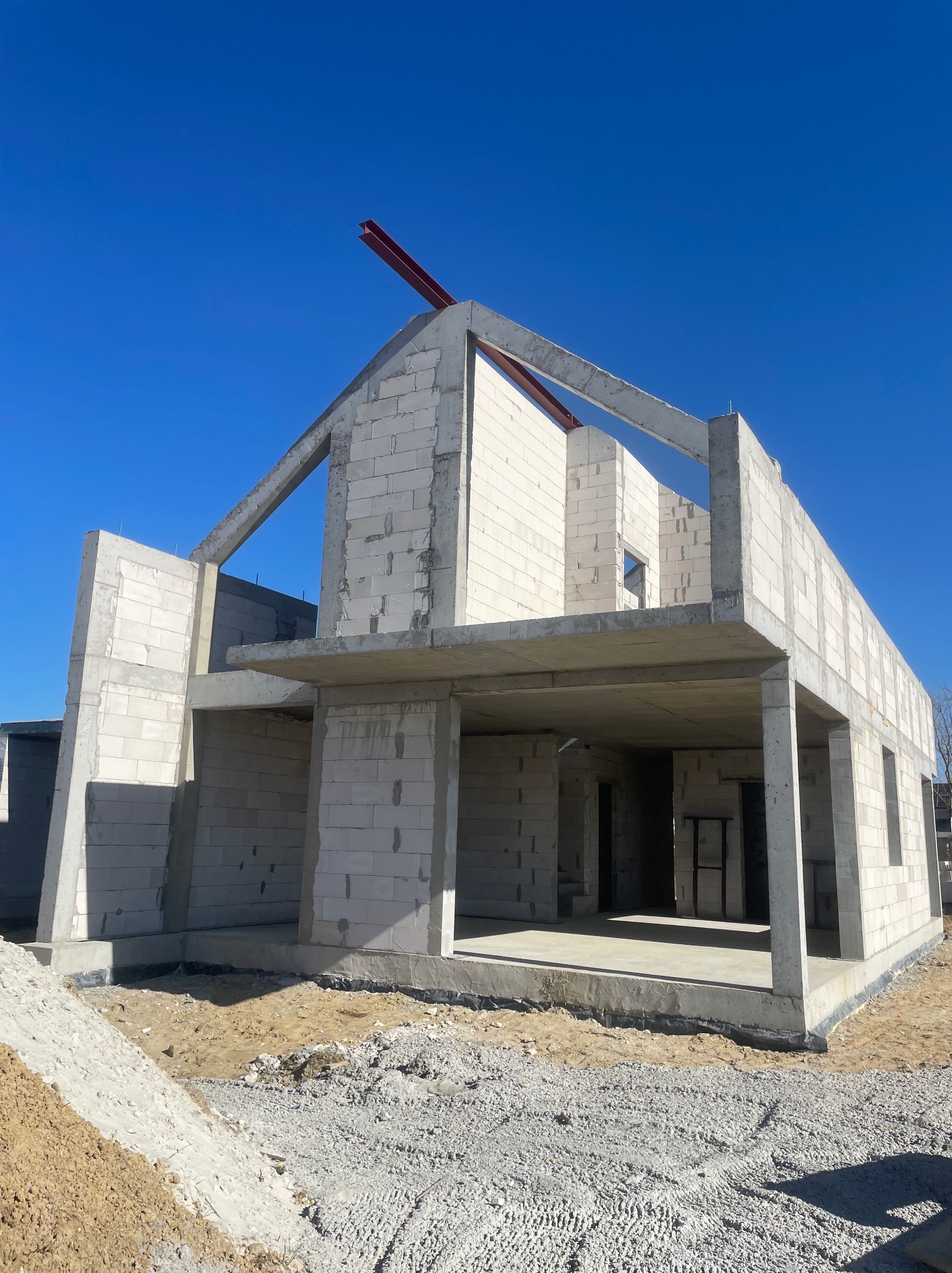 Niedokończony dwukondygnacyjny dom z białych bloczków, z widoczną czerwoną belką na dachu, w surowym stanie, na tle błękitnego nieba.
