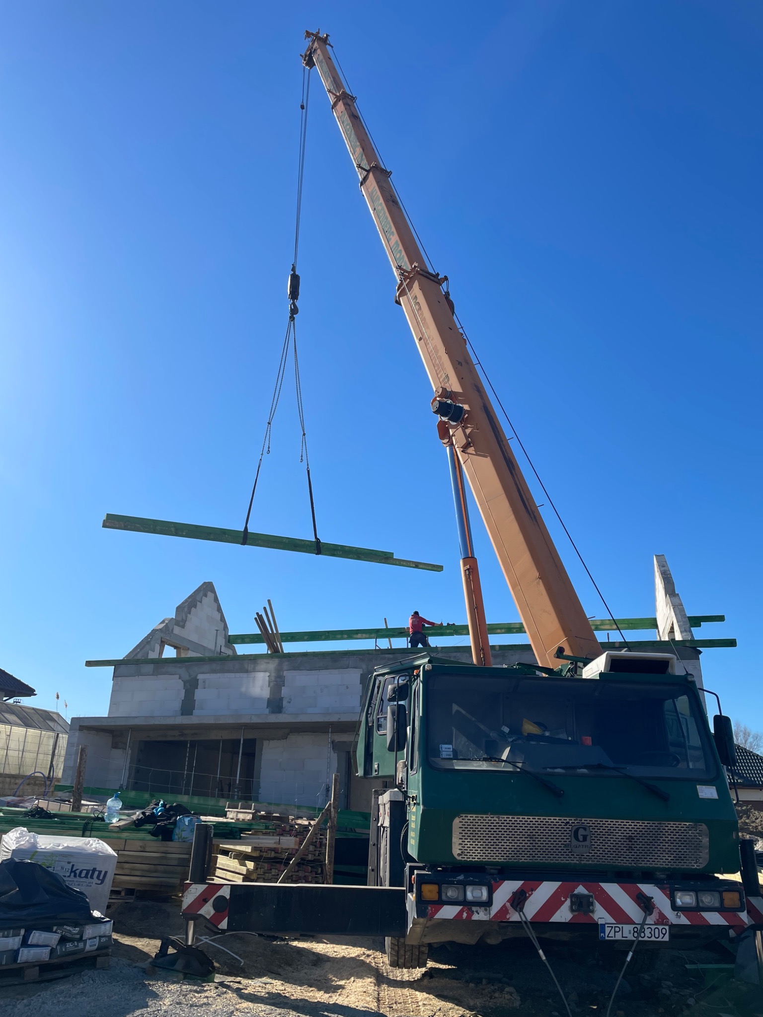 Żuraw budowlany podnosi belkę na budowę domu z białego pustaka. Widoczny pracownik na dachu i materiały budowlane. Słoneczny dzień, niebieskie niebo.