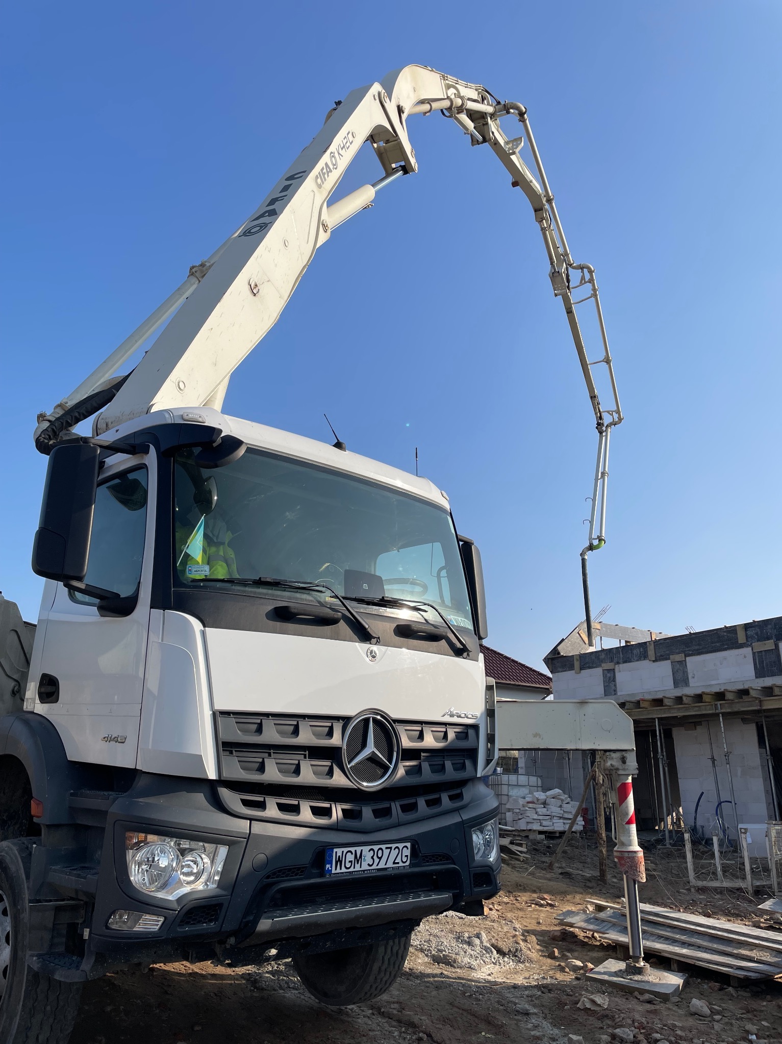 Betonomieszarka marki Mercedes na budowie domu jednorodzinnego z białych bloczków, ramię pompy skierowane w stronę budynku.