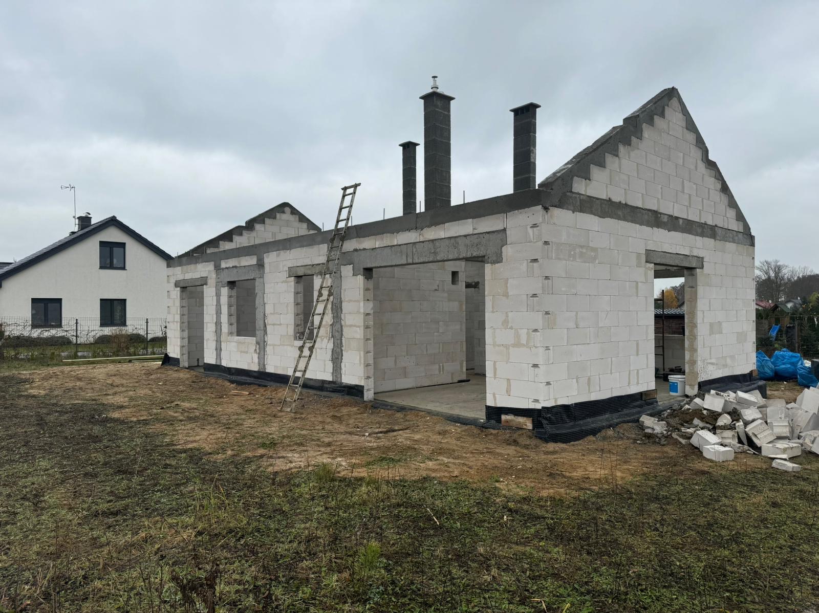Surowy stan domu z bloczków betonu komórkowego, z widocznymi kominami i drewnianą drabiną opartą o ścianę, na tle pochmurnego nieba i sąsiedniego domu.