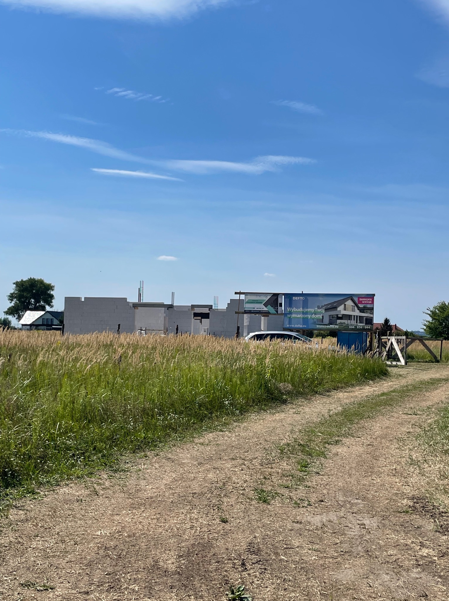 Budowa domu z białych bloczków, widoczny billboard z wizualizacją gotowego projektu. Polna droga prowadzi do placu budowy, wysoka trawa na pierwszym planie.